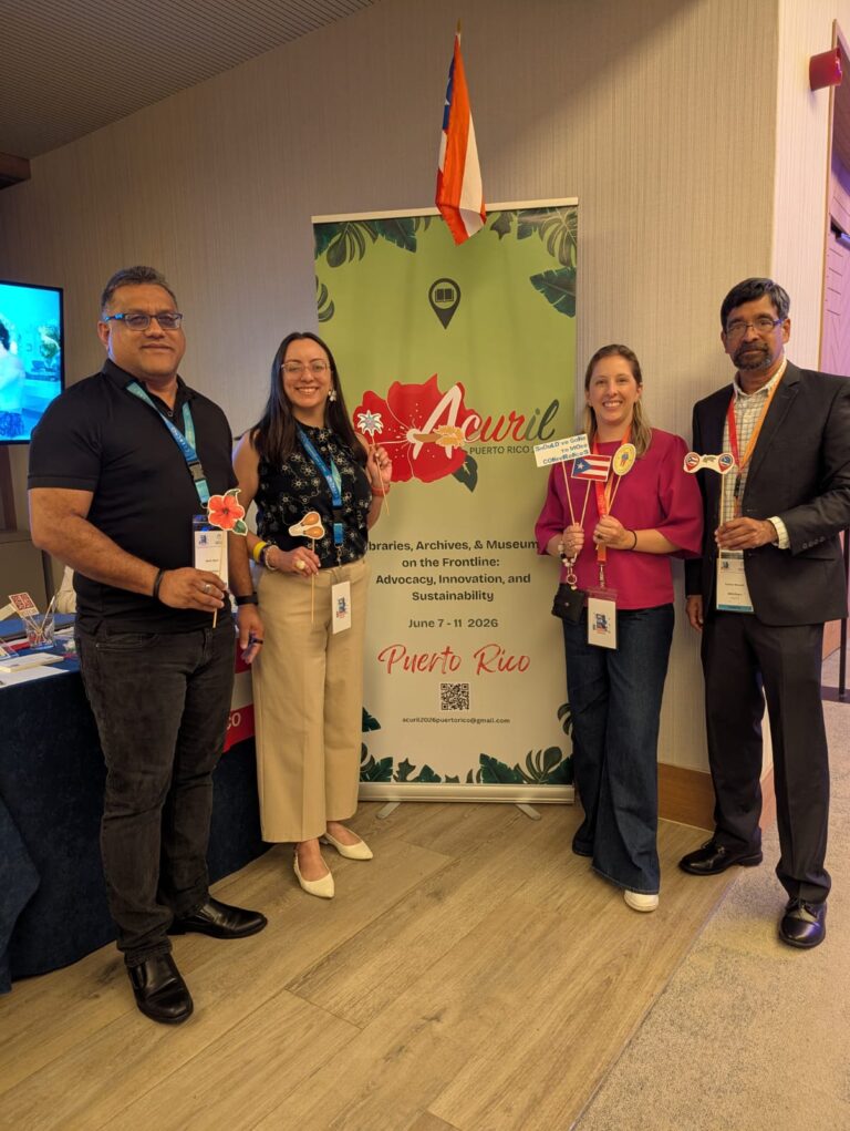 El equipo combinado de ACURIL, Bibliotheca y ECCA posando con banderas en su stand para la conferencia de 2026 en Puerto Rico.