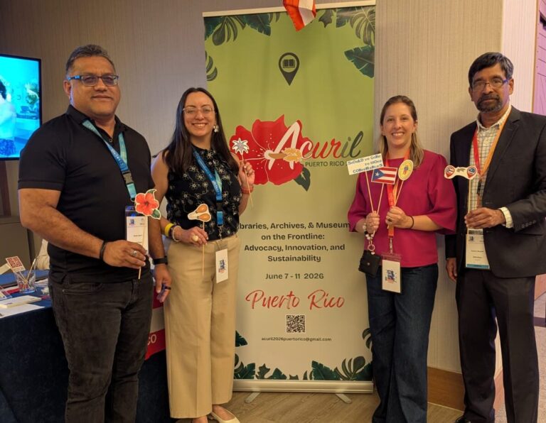 El equipo combinado de ACURIL, Bibliotheca y ECCA posando con banderas en su stand para la conferencia de 2026 en Puerto Rico.